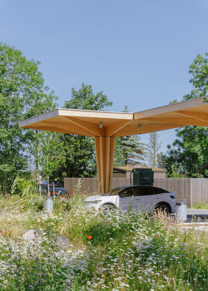 Clever lynladestation i træ med en elbil under taget, omgivet af vilde blomster